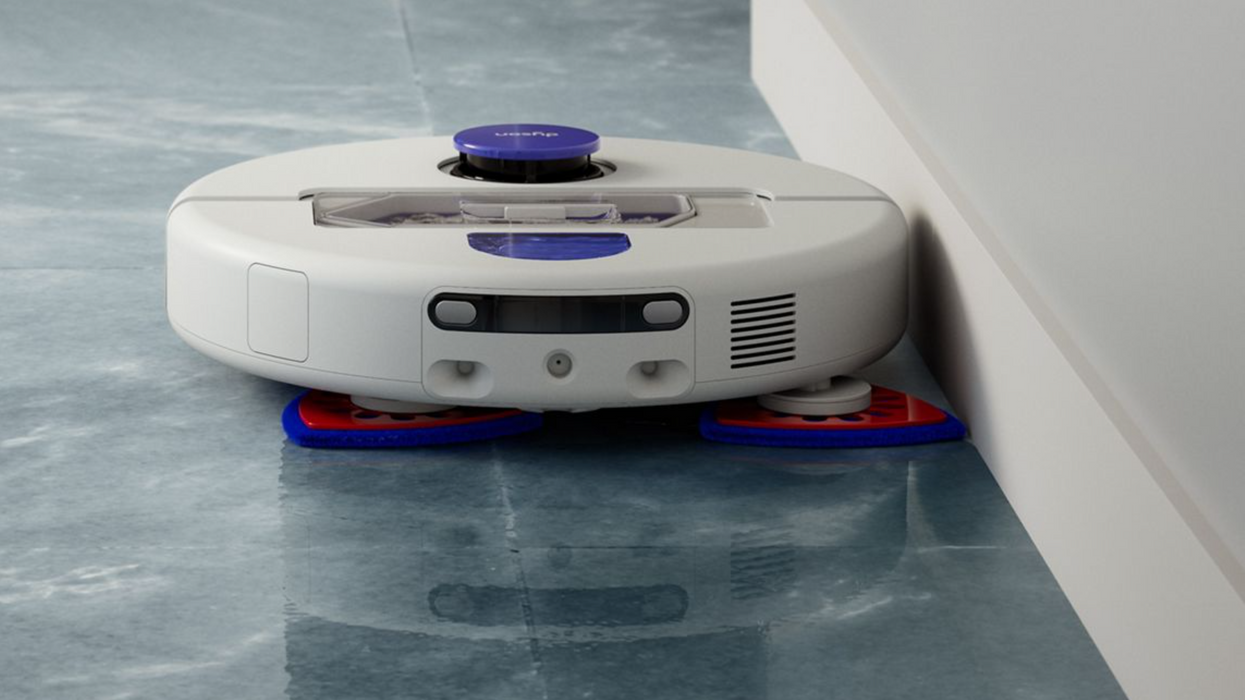 A white robotic floor cleaner with purple and blue accents, shown on a glossy blue-gray floor against a white wall corner.