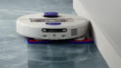 A white robotic floor cleaner with purple and blue accents, shown on a glossy blue-gray floor against a white wall corner.