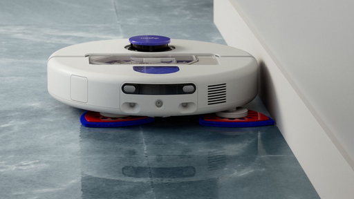 A white robotic floor cleaner with purple and blue accents, shown on a glossy blue-gray floor against a white wall corner.