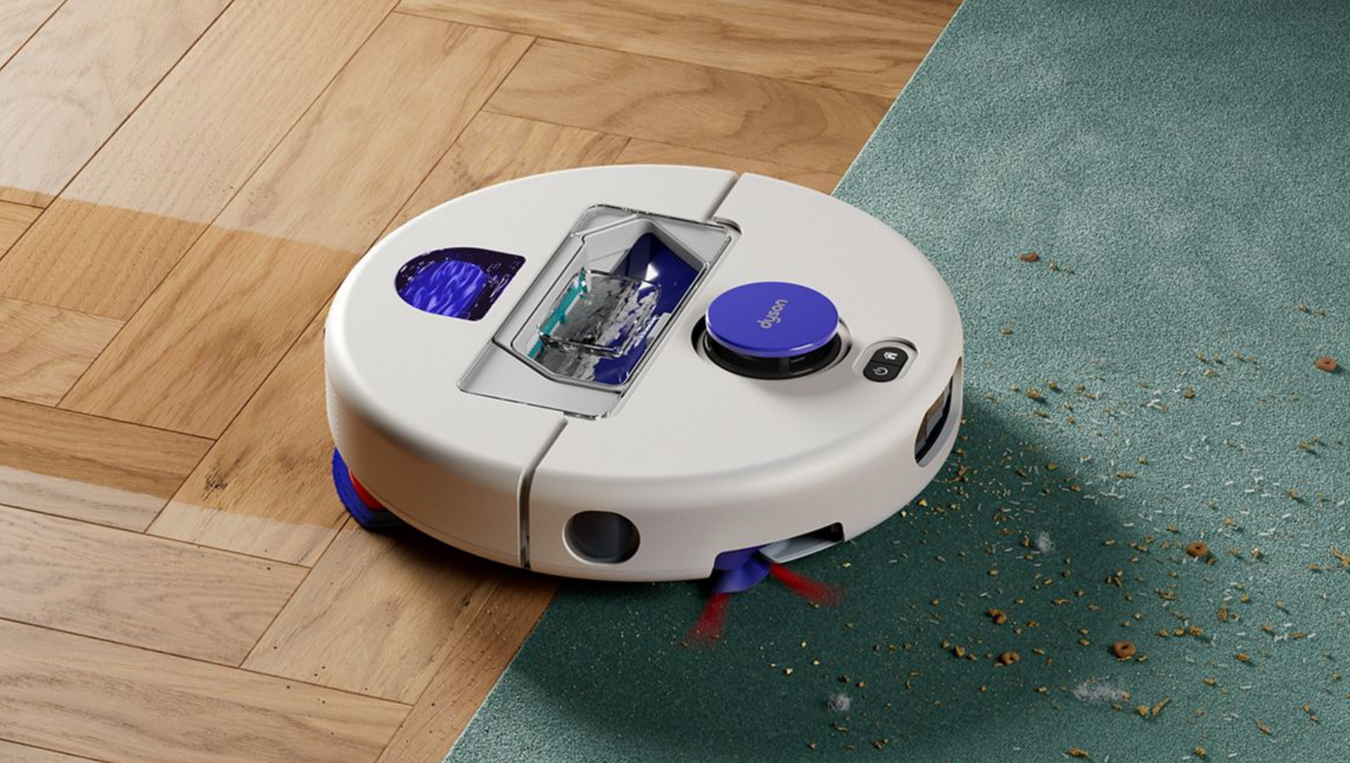 A round white robotic vacuum cleaner with blue accents is shown on a split hardwood and green carpet floor, with debris visible on the carpet.