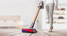 A cordless stick vacuum cleaner with a metallic copper-orange handle and purple, gray, and red head is being used by a person in light gray leggings on a light wood floor with crumbs. The background is a softly blurred modern room.
