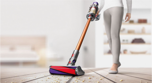 A cordless stick vacuum cleaner with a metallic copper-orange handle and purple, gray, and red head is being used by a person in light gray leggings on a light wood floor with crumbs. The background is a softly blurred modern room.