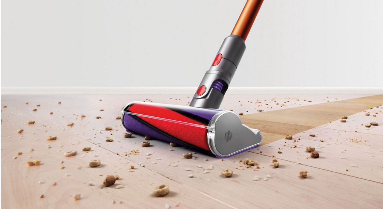 A cordless stick vacuum cleaner head with a red, purple, and silver design, shown cleaning up crumbs and debris on a light-colored wooden floor.