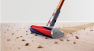 A cordless stick vacuum cleaner head with a red, purple, and silver design, shown cleaning up crumbs and debris on a light-colored wooden floor.