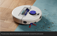 A round robotic vacuum cleaner shown on a split hardwood and blue carpet floor, with debris being picked up and a text overlay at the bottom describing its dust sensor and suction power.