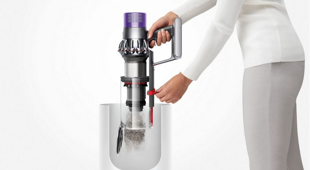 A person in a white ribbed sweater and light gray pants holds a cordless vacuum cleaner over a clear container, with dust falling into the container. The background is a clean white studio backdrop.