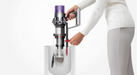A person in a white ribbed sweater and light gray pants holds a cordless vacuum cleaner over a clear container, with dust falling into the container. The background is a clean white studio backdrop.