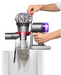 A cordless vacuum cleaner with a transparent dust bin and purple and silver body, shown with a person's hands demonstrating how to empty it into a white container against a white background.

