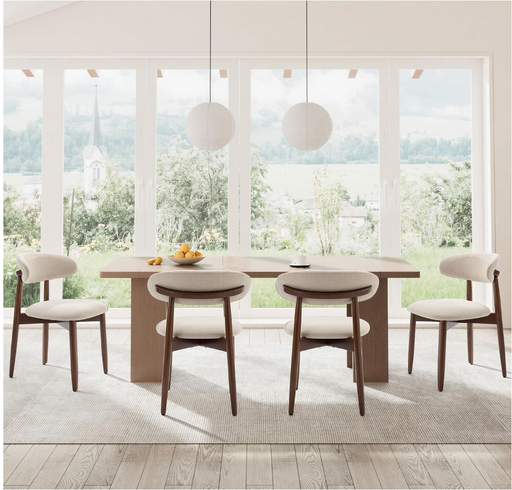 A modern dining room set featuring a rectangular light wood table with a thick base and four matching chairs with light beige upholstered seats and backs, set on a textured rug in front of large windows with a scenic view.