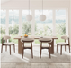 A modern dining room set featuring a rectangular light wood table with a thick base and four matching chairs with light beige upholstered seats and backs, set on a textured rug in front of large windows with a scenic view.