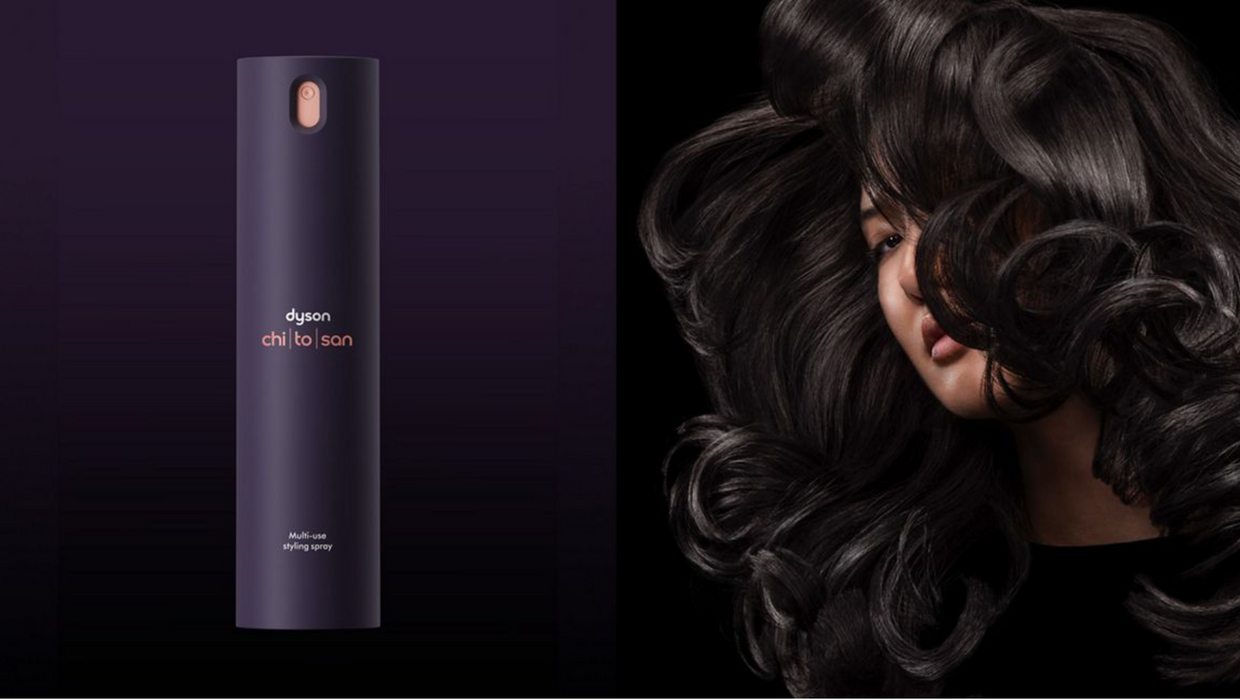A tall cylindrical Dyson chiller ion hair care bottle in dark purple stands on a dark purple background, next to a model with long, glossy, dark brown to black wavy hair.