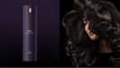 A tall cylindrical Dyson chiller ion hair care bottle in dark purple stands on a dark purple background, next to a model with long, glossy, dark brown to black wavy hair.