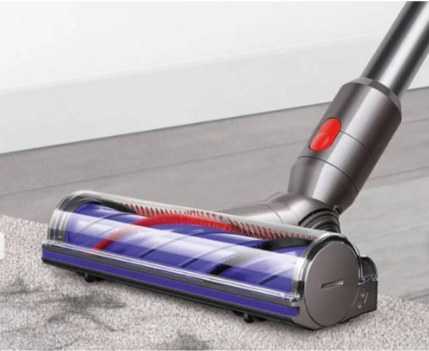 A close-up of a vacuum cleaner head attachment moving across a gray carpet in front of a white baseboard and wall.
