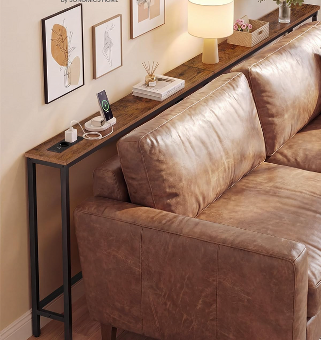 A narrow console table with a rustic dark wood top and black metal legs, styled next to a brown leather sofa in a warm living room setting.