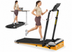 A woman is shown running on a treadmill in the foreground, with a smaller inset showing another woman using a standing desk attachment on the treadmill bed. The treadmill is black with a bright orange base panel and gray side housing, photographed on a clean white background.